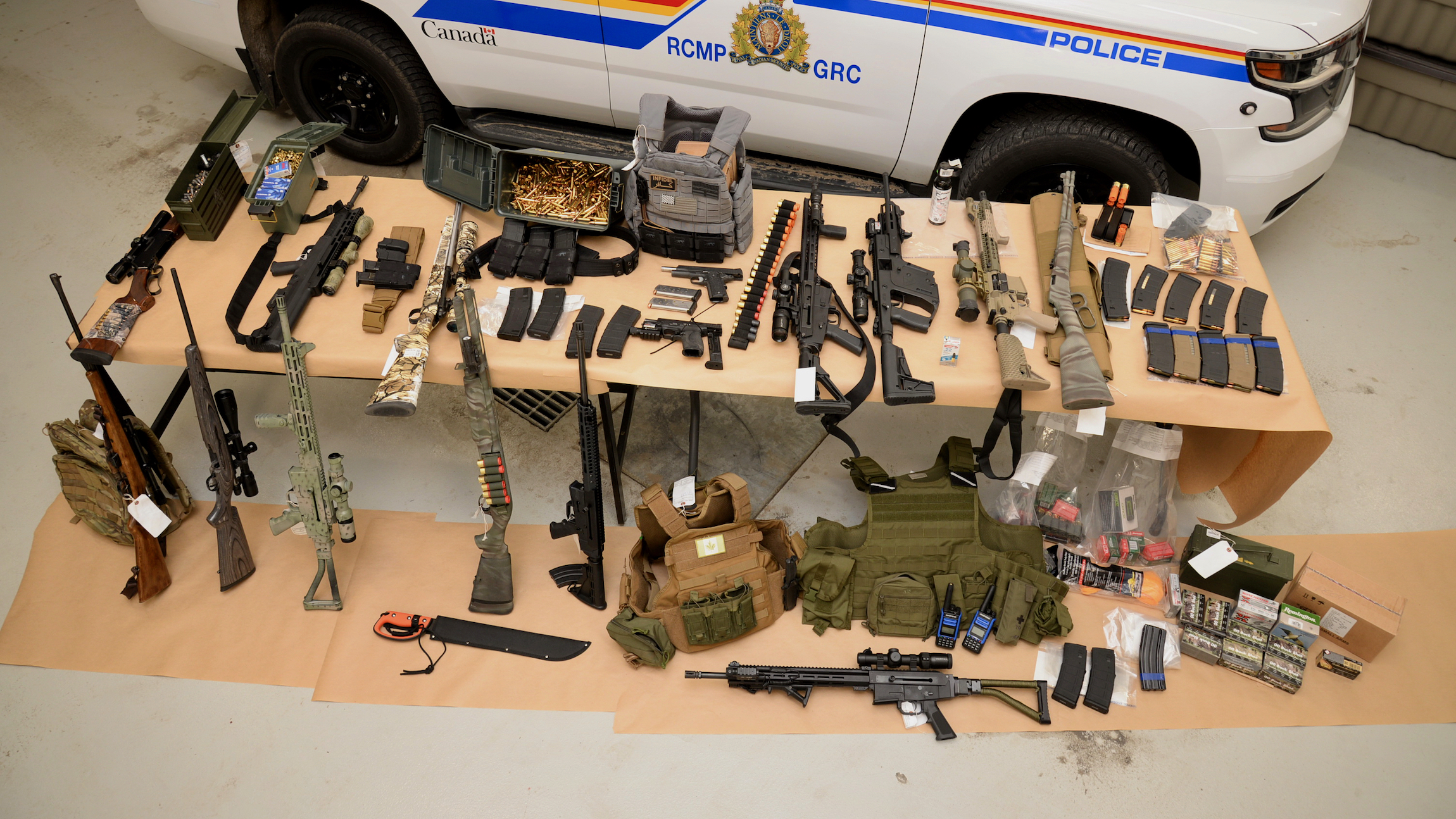 Long guns, body armor, ammunition are laid out on a table with a Royal Canadian mounted police car in the background