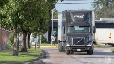 A brown tractor-trailer with the logo of TForce Freight travels on a city street with a trailer of UPS seen in the background.