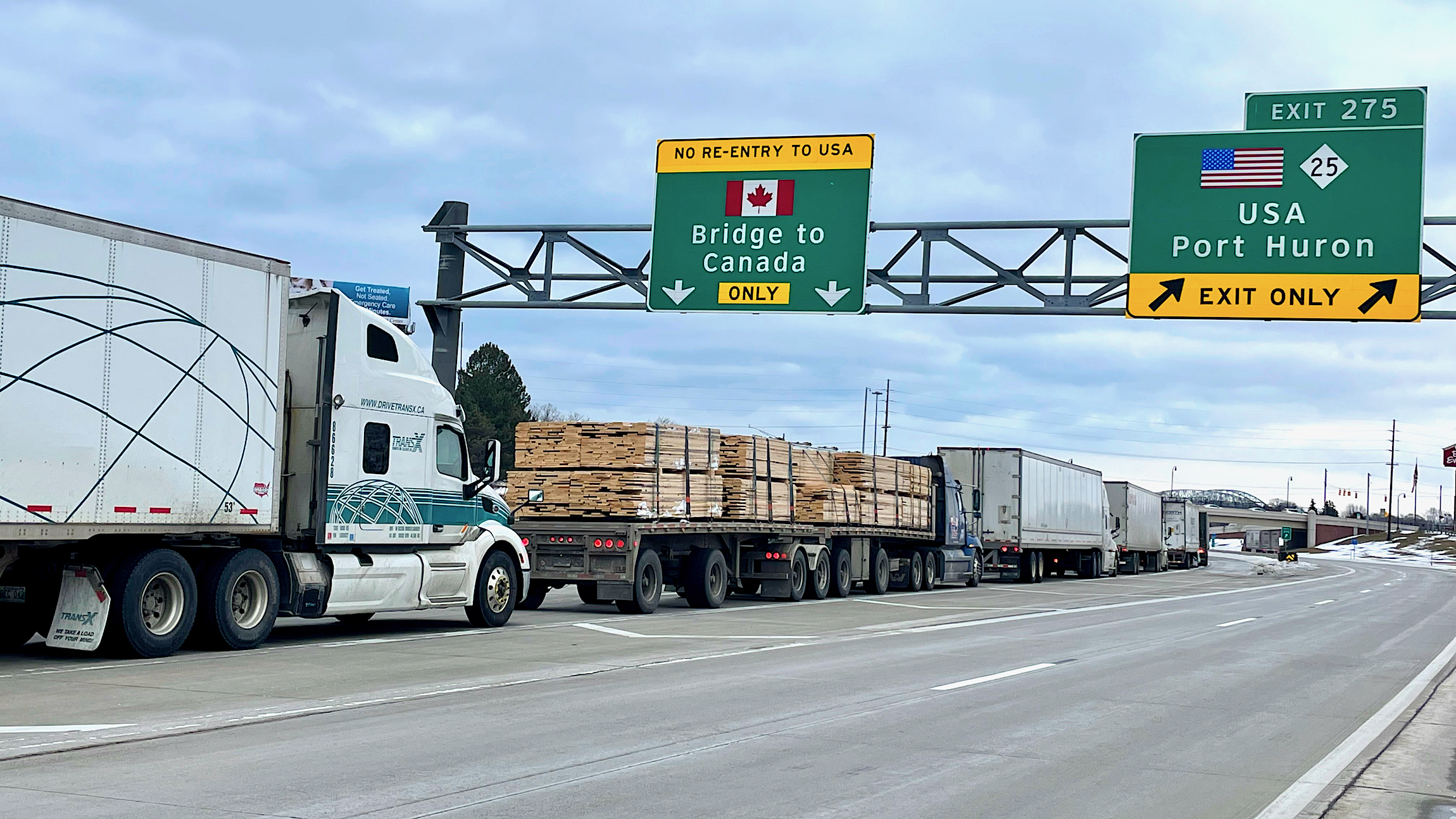 A line of trucks move on a road where a sign reads bridge to Canada