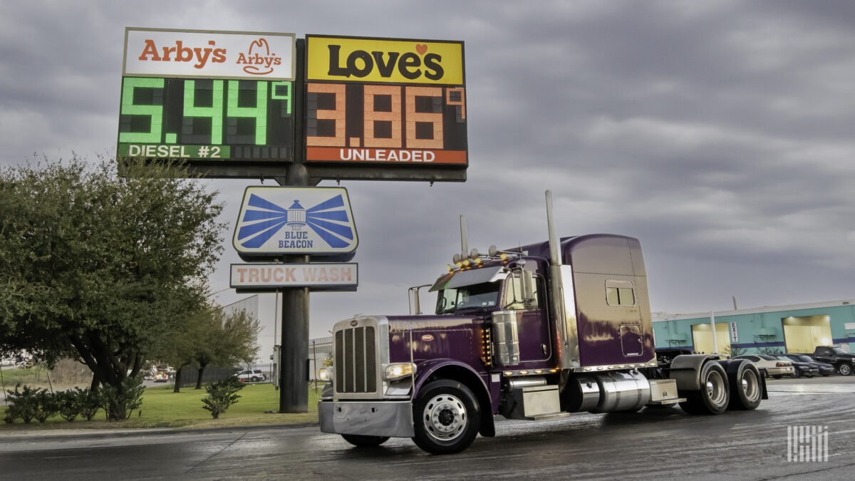 A red semi-truck is parked in front of a sign displaying the prices of diesel and regular gas