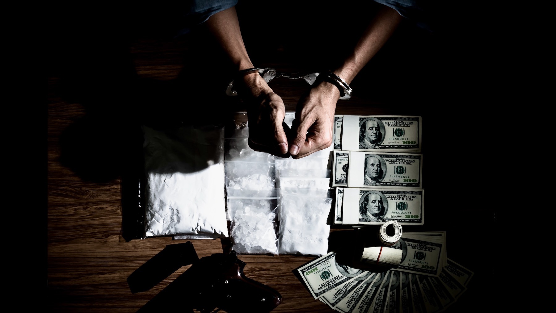 A person is handcuffed at at a table with drugs, a gun and money