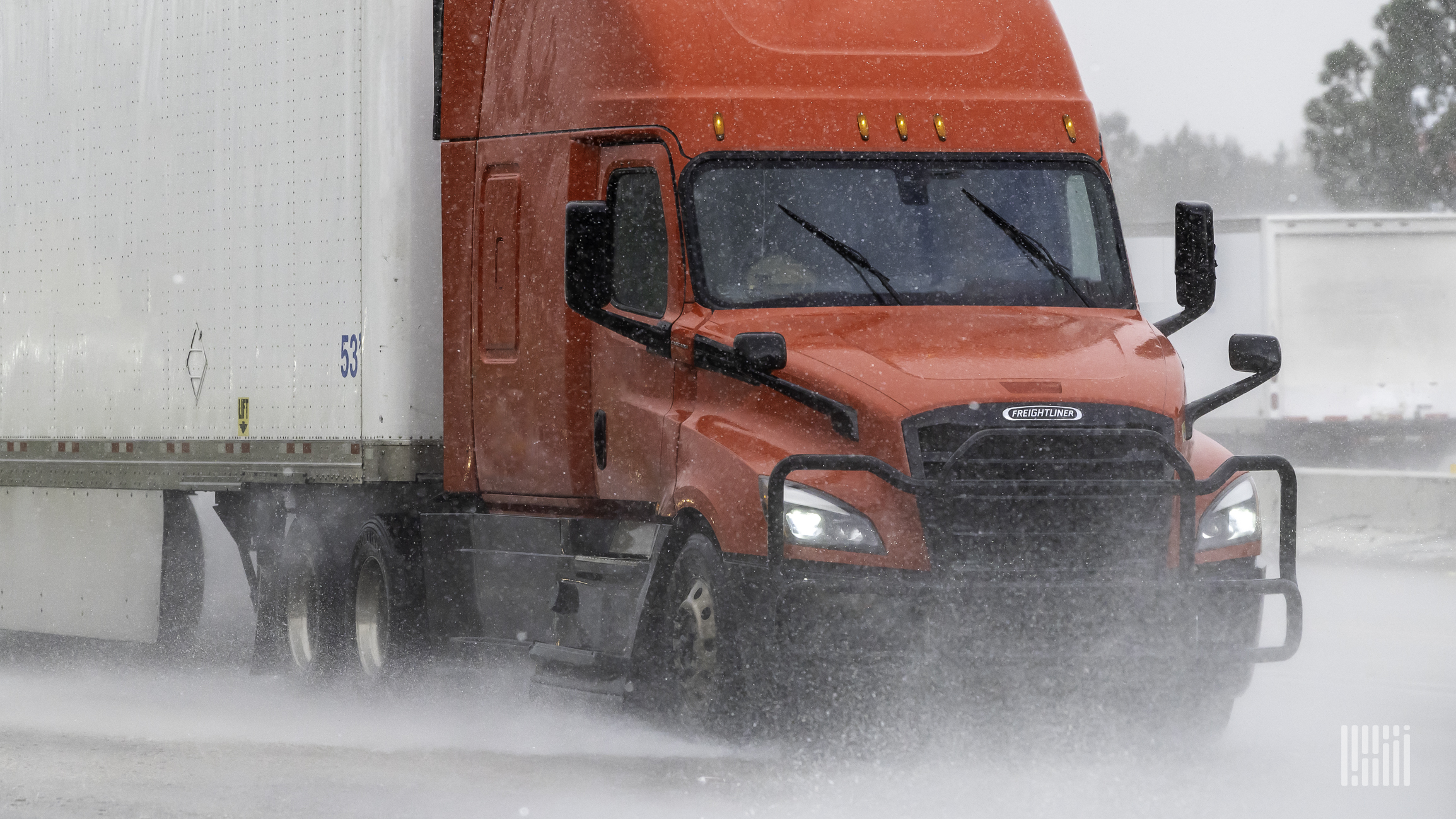 A red semi truck with a white trailer drives through rain