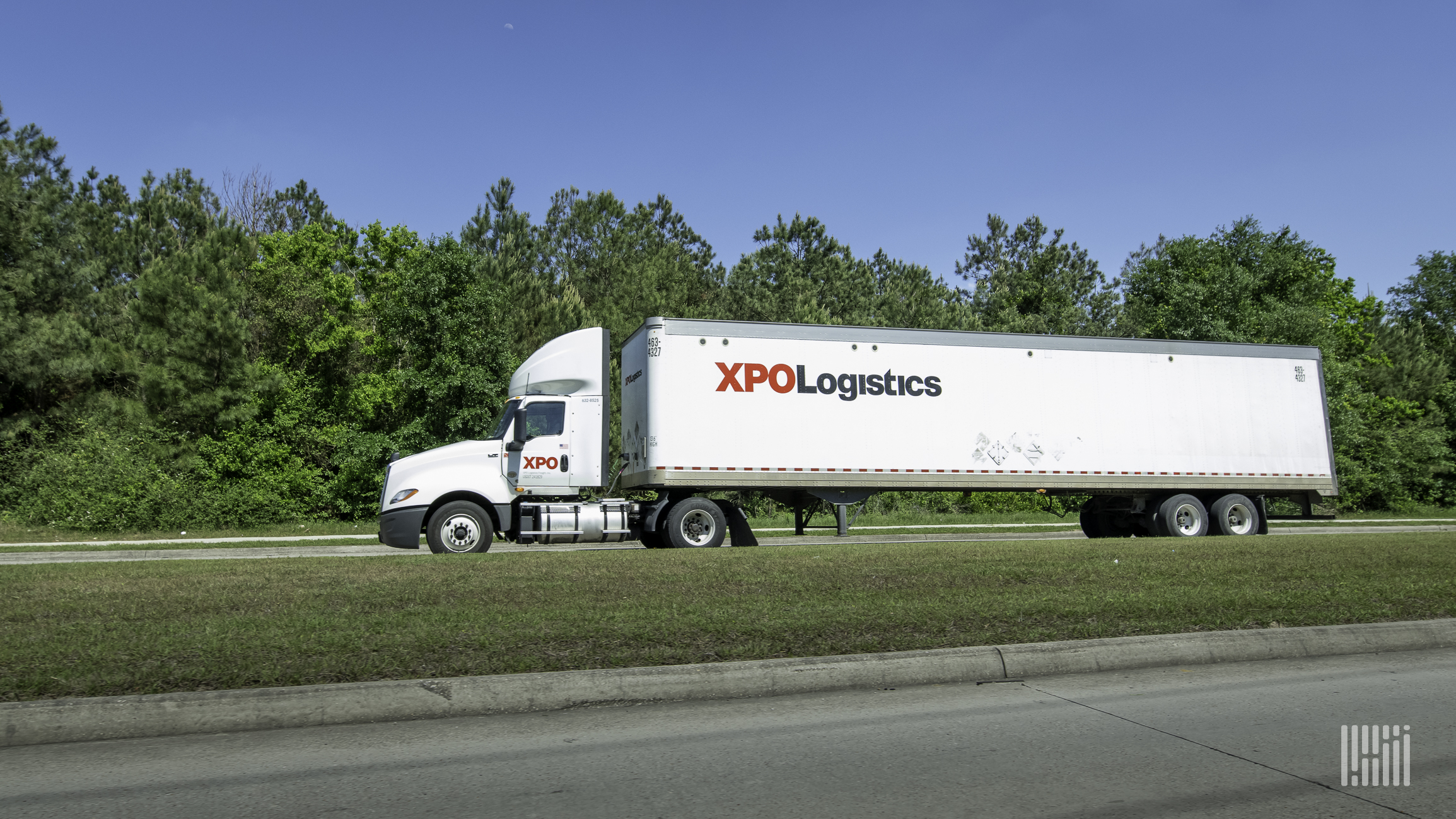 A white XPO Logistics tractor-trailer travels on a major road with a green landscape and blue skies in the background.