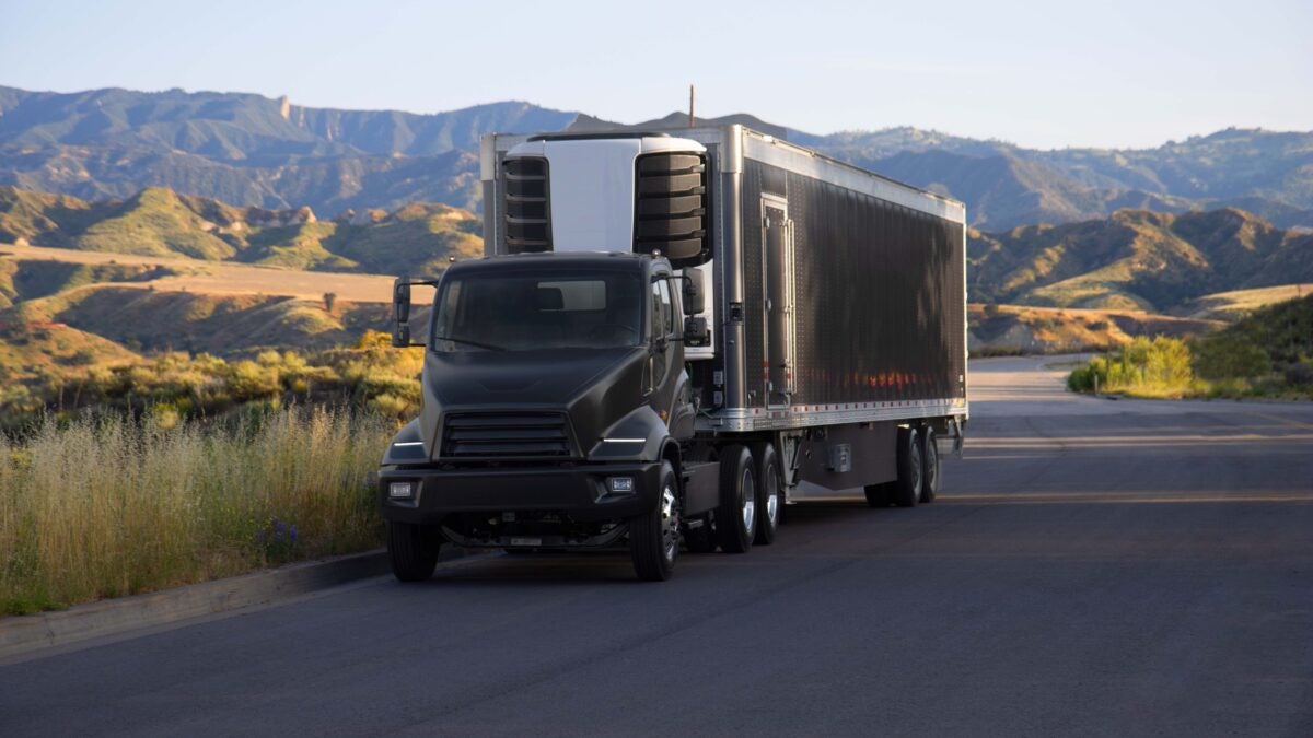 A black Xos Class 8 refrigerated semi-truck on a desert highway framed by mountains.