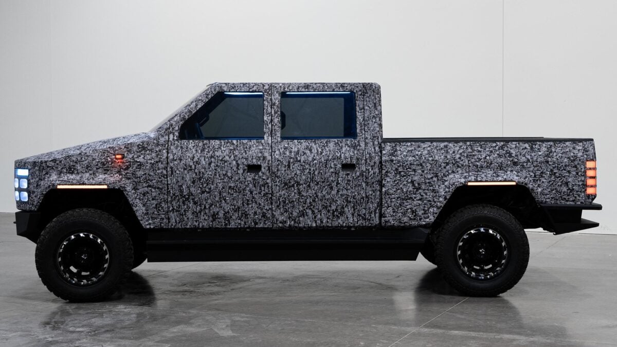 A camouflaged side view of the Atlis commercial pickup truck prototype with front and rear lights illuminated.