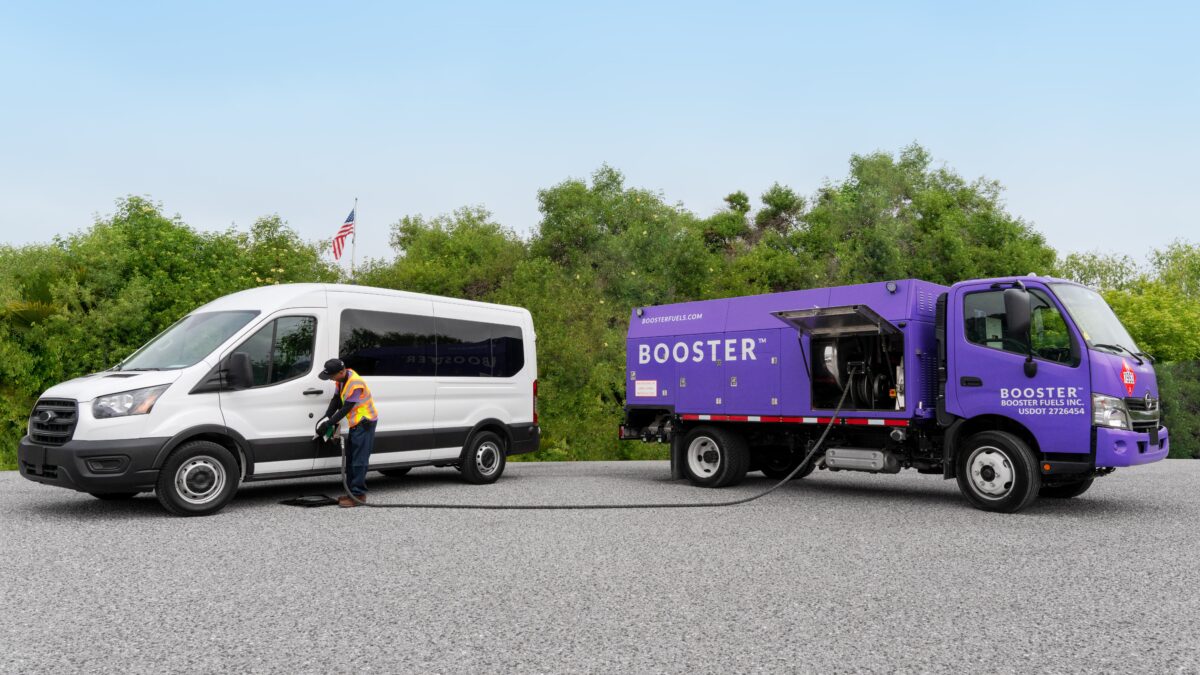 A Booster employee wearing a yellow and orange safety vest fills a white Ford Transit van with fuel from a purple Booster mobile fuel truck.