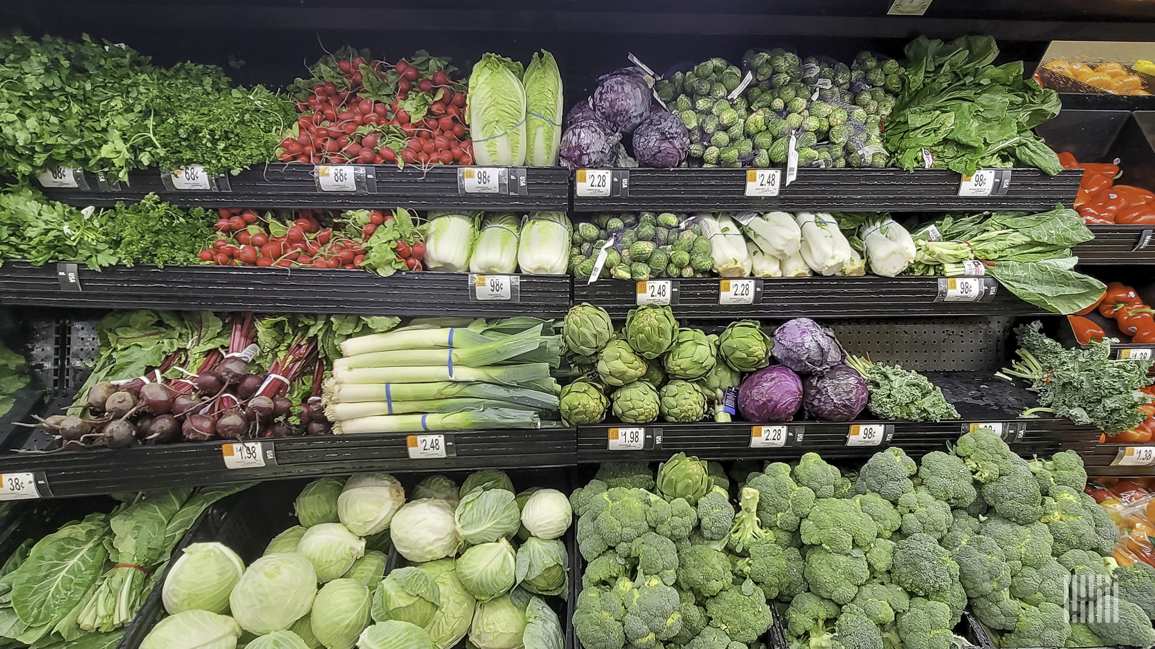 A produce section of a grocery store shows foods such as broccoli, celery, cabbage, radishes, and lettuce.