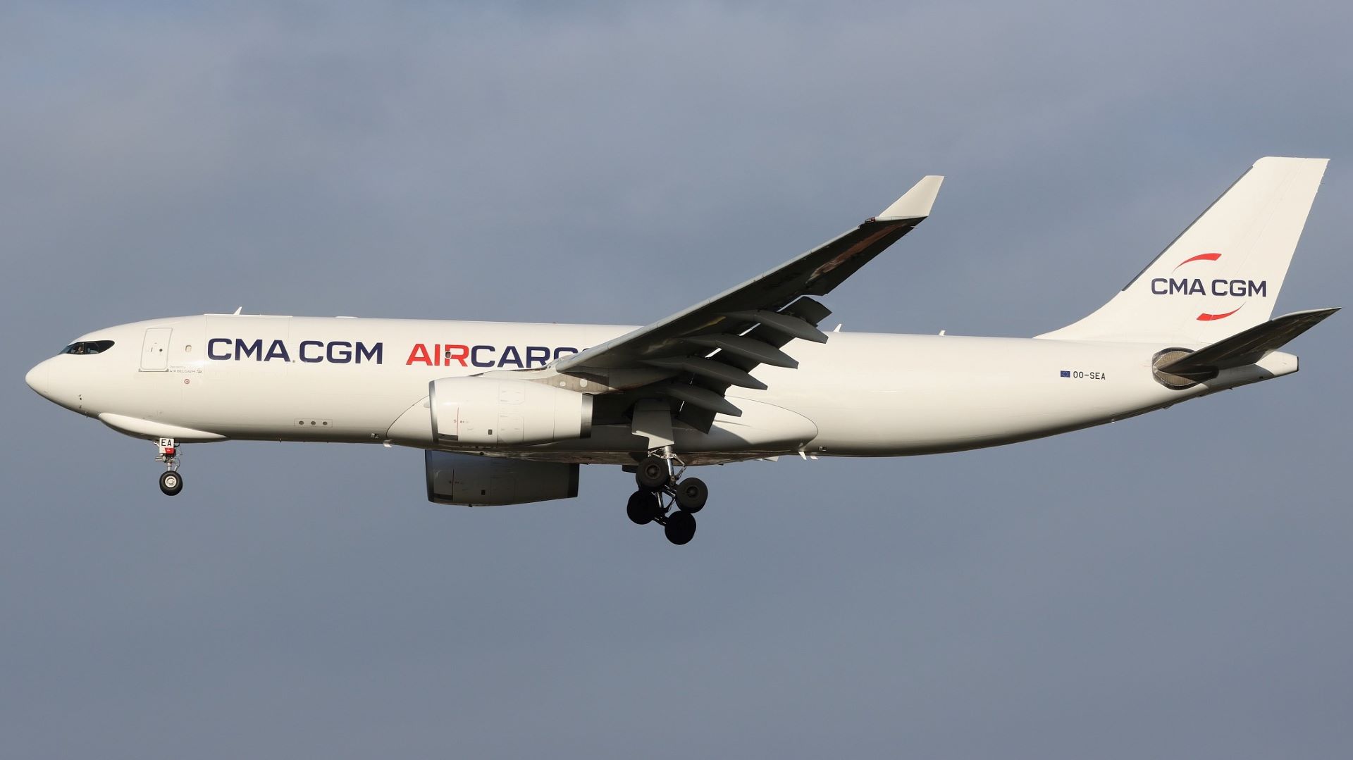 A white CMA CGM Air Cargo jet comes in for landing with wheels down, view from below.