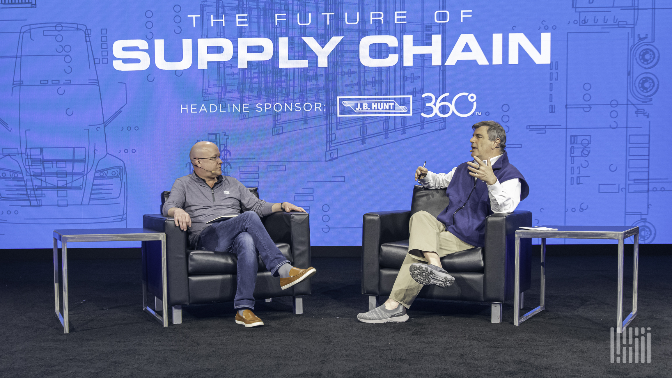 Two men sitting in chairs on a stage and talking to each other.