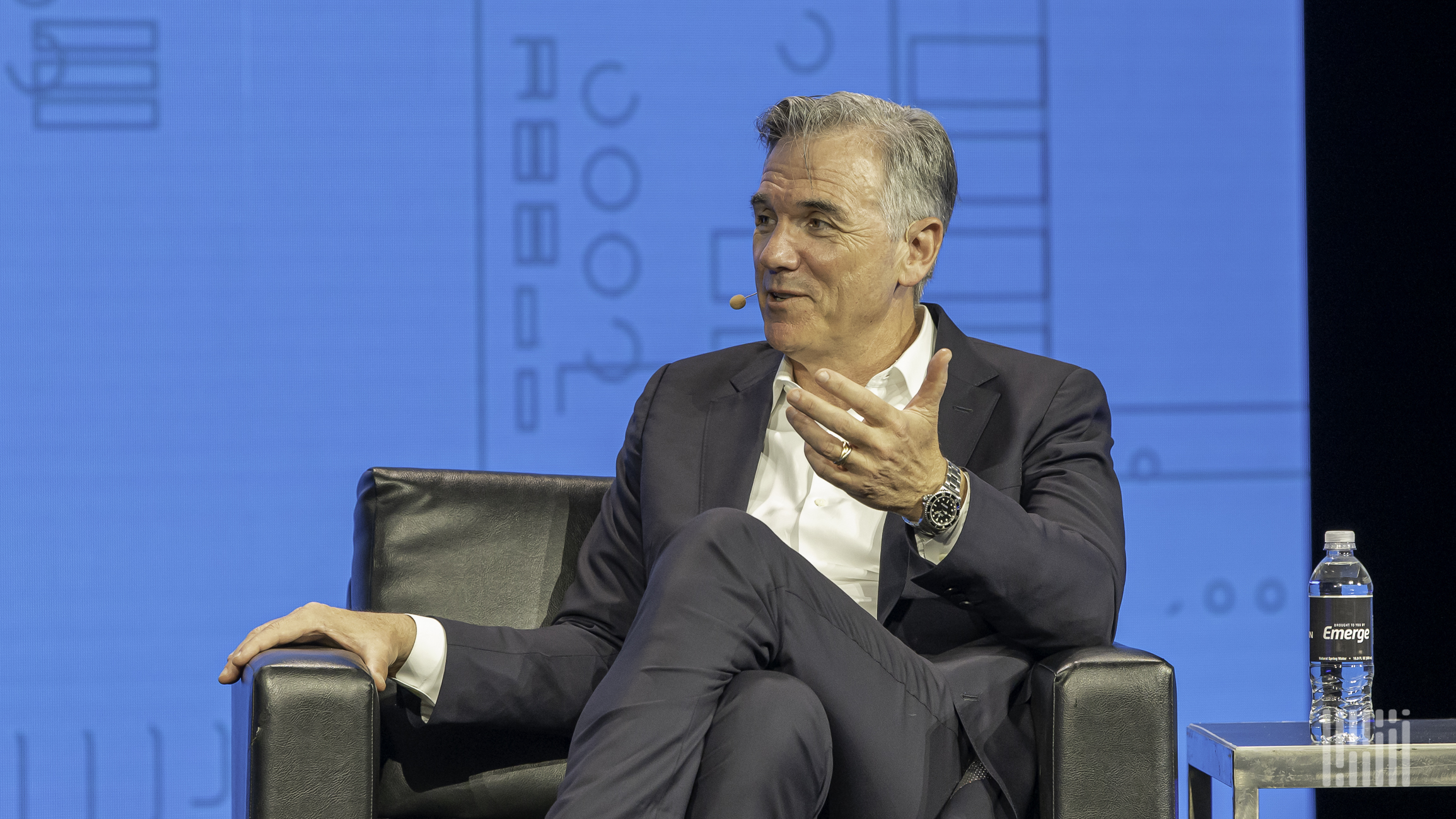 A man sitting on a leather chair talks to another person with one arm extended.