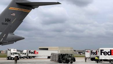 A tail of a military plane and FedEx trucks shown on an airport tarmac as a tractor moves pallets.