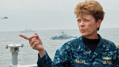 A woman in a blue Navy camouflage uniform with the name Phillips sewn on it, points while on the deck of a ship,