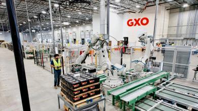 A man works in a warehouse alongside robots while the logo of the company GXO Logistics is visible
