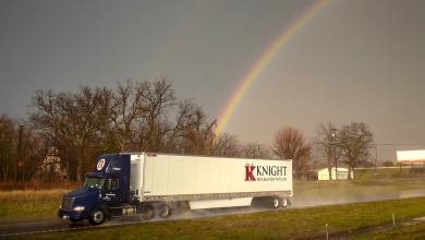 A tractor-trailer with the logo of Knight Transportation drives on a road with storm clouds and a rainbow behind it, illustrating a downturn in the trucking industry.