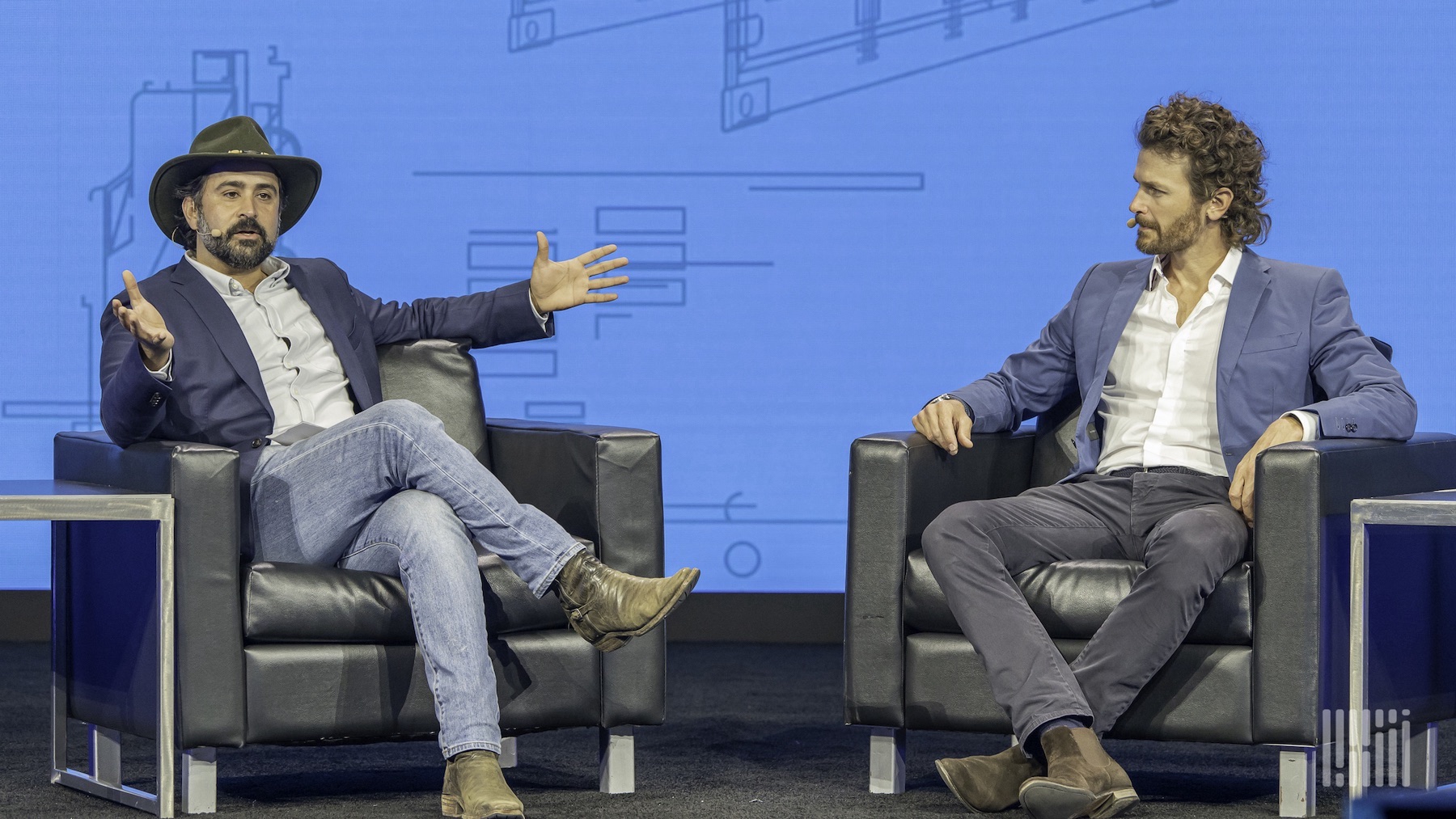 Two men sitting in leather chairs talking, as the one on the left gestures with his arms out.