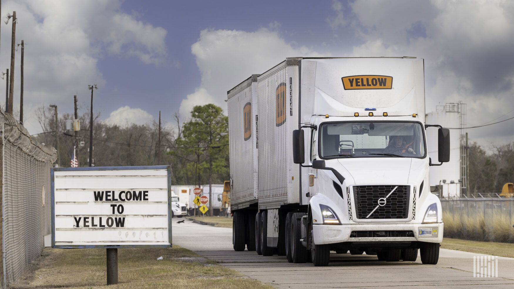 A semi truck with a double trailer with the logo of the company Yellow driving on a road next to a sign reading Welcome to Yellow