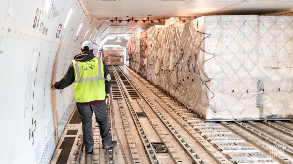 Man in yellow vest monitors loading of pallets on roller system inside body of aircraft.