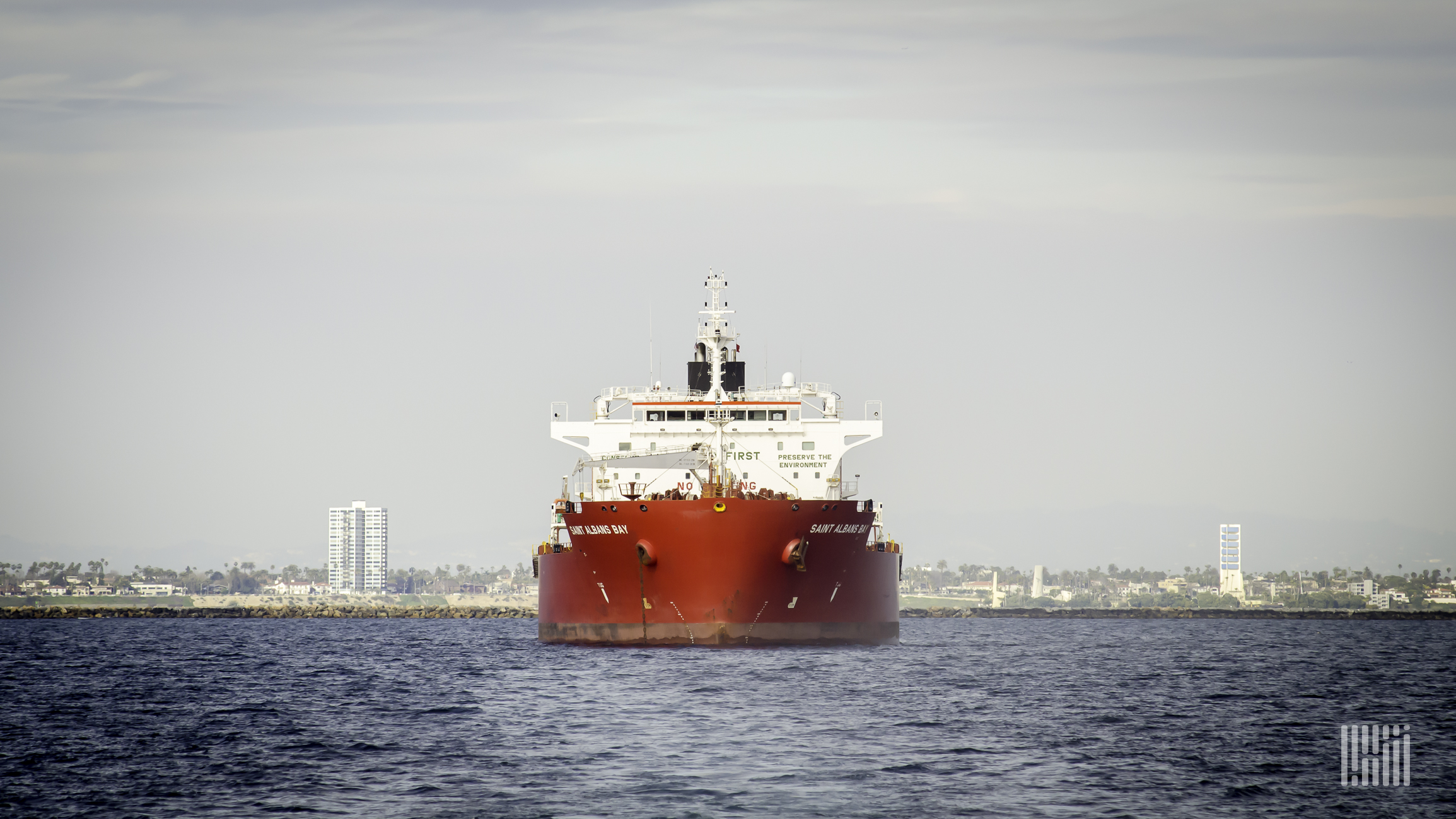 A red and white container ship is traveling on the ocean with the shoreline in the background.