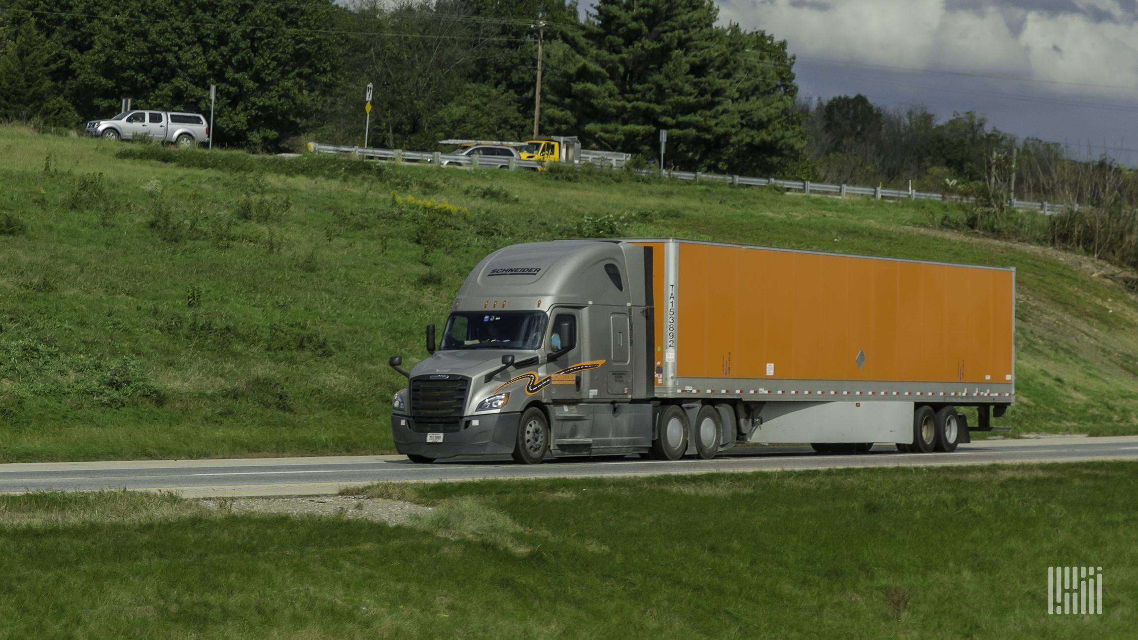 A grey and orange Schneider truck travels down a road green trees and grass in the background.