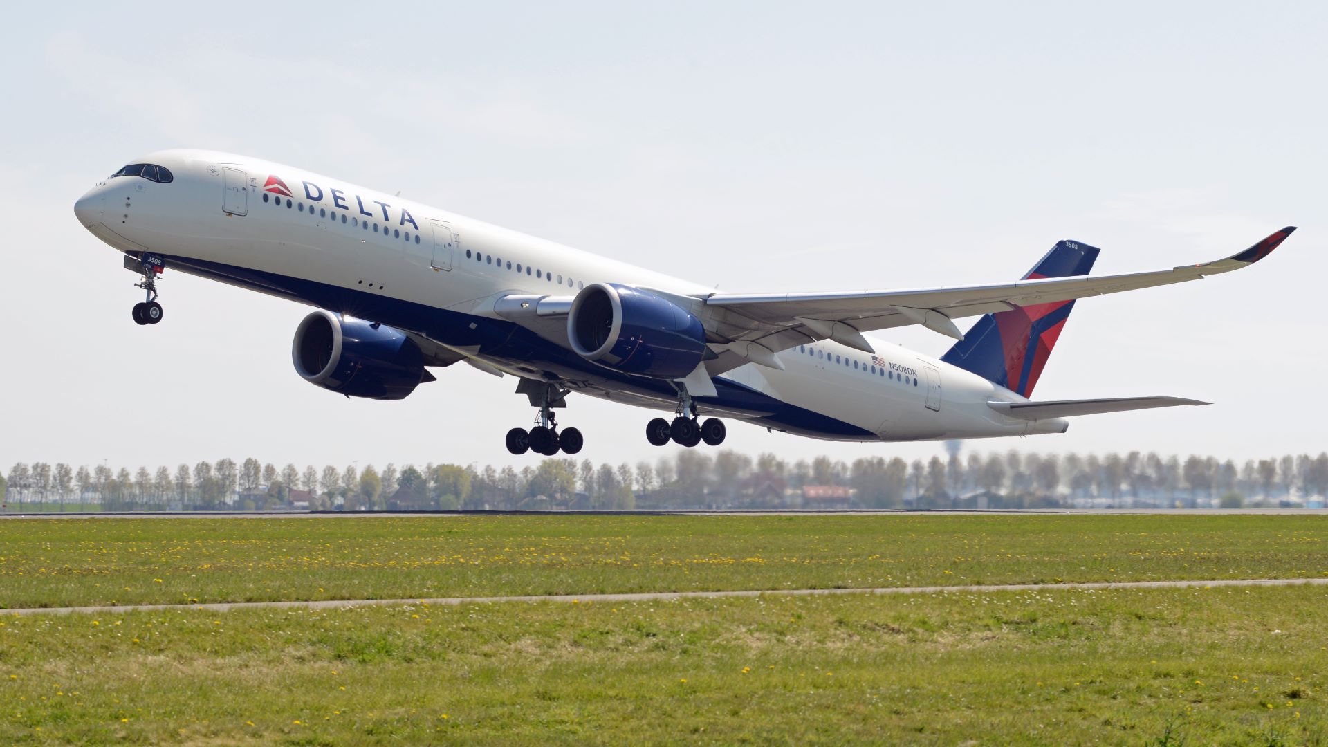 A large white jet with blue tail and Delta logo takes off from a grassy airport.