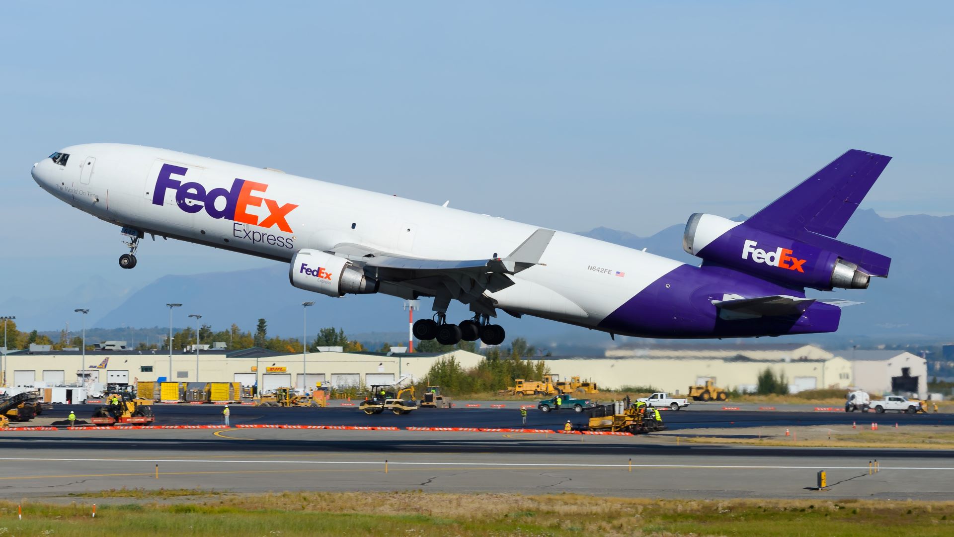 A white FedEx jet with a blue tail lifts off the runway with warehouses in the background.
