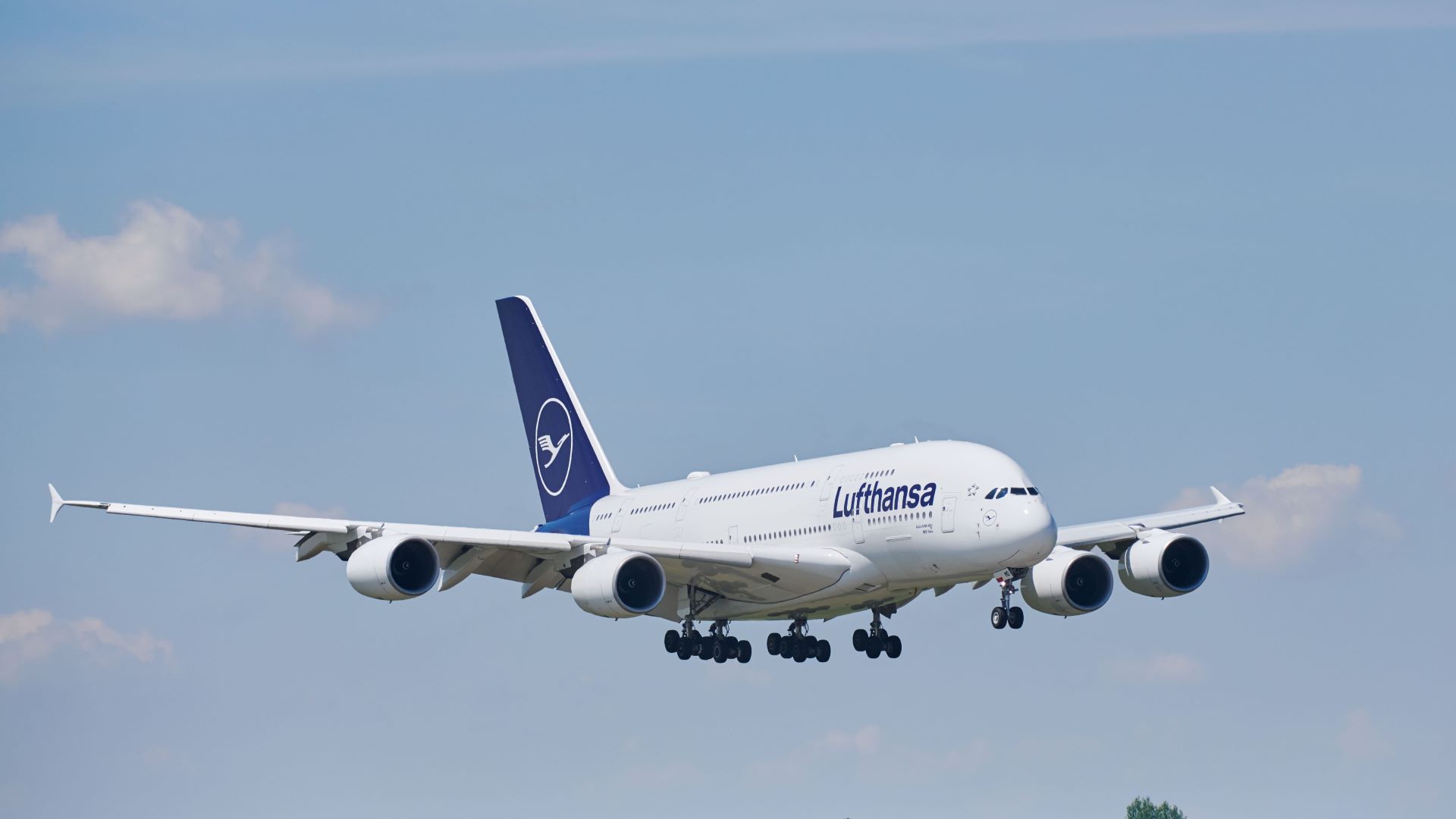 A large white A380 jet with blue tail and Lufthansa logo comes in for landing with wheels down against a blue sky.