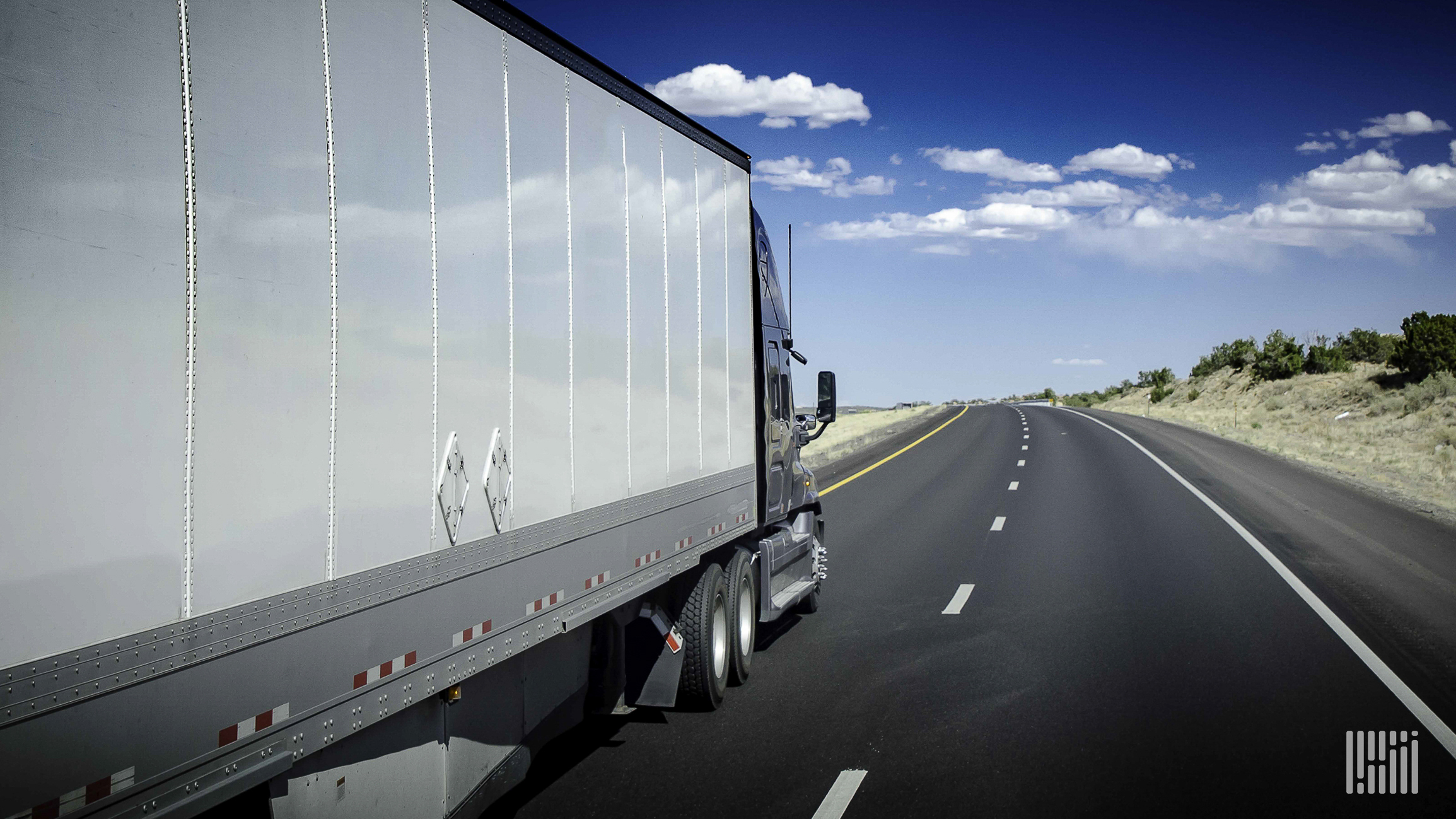 A white semi-truck is traveling down a road with a blue sky in the background.