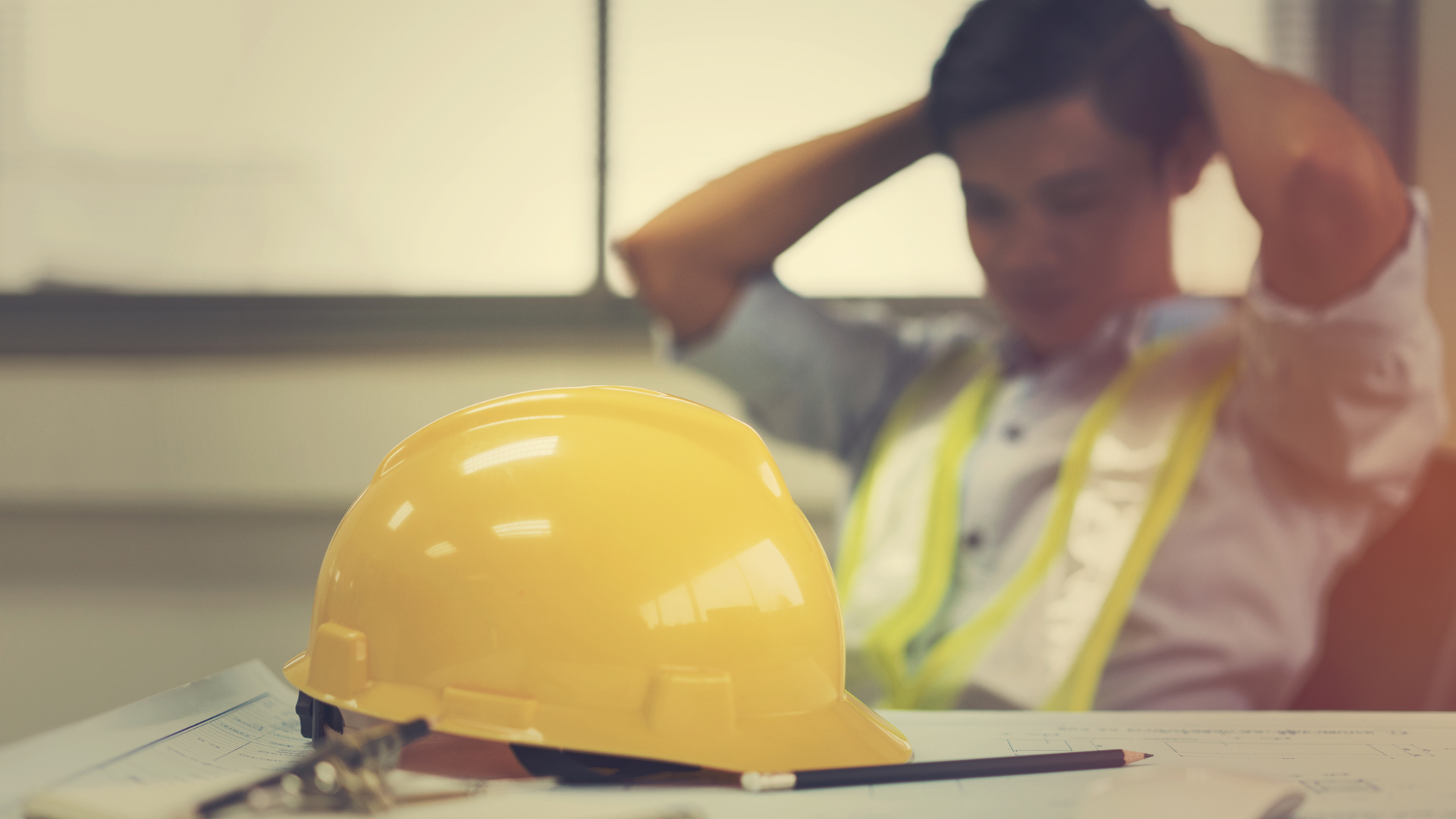 A worker sitting down with his hands behind his head. In front of him is a hard hat.
