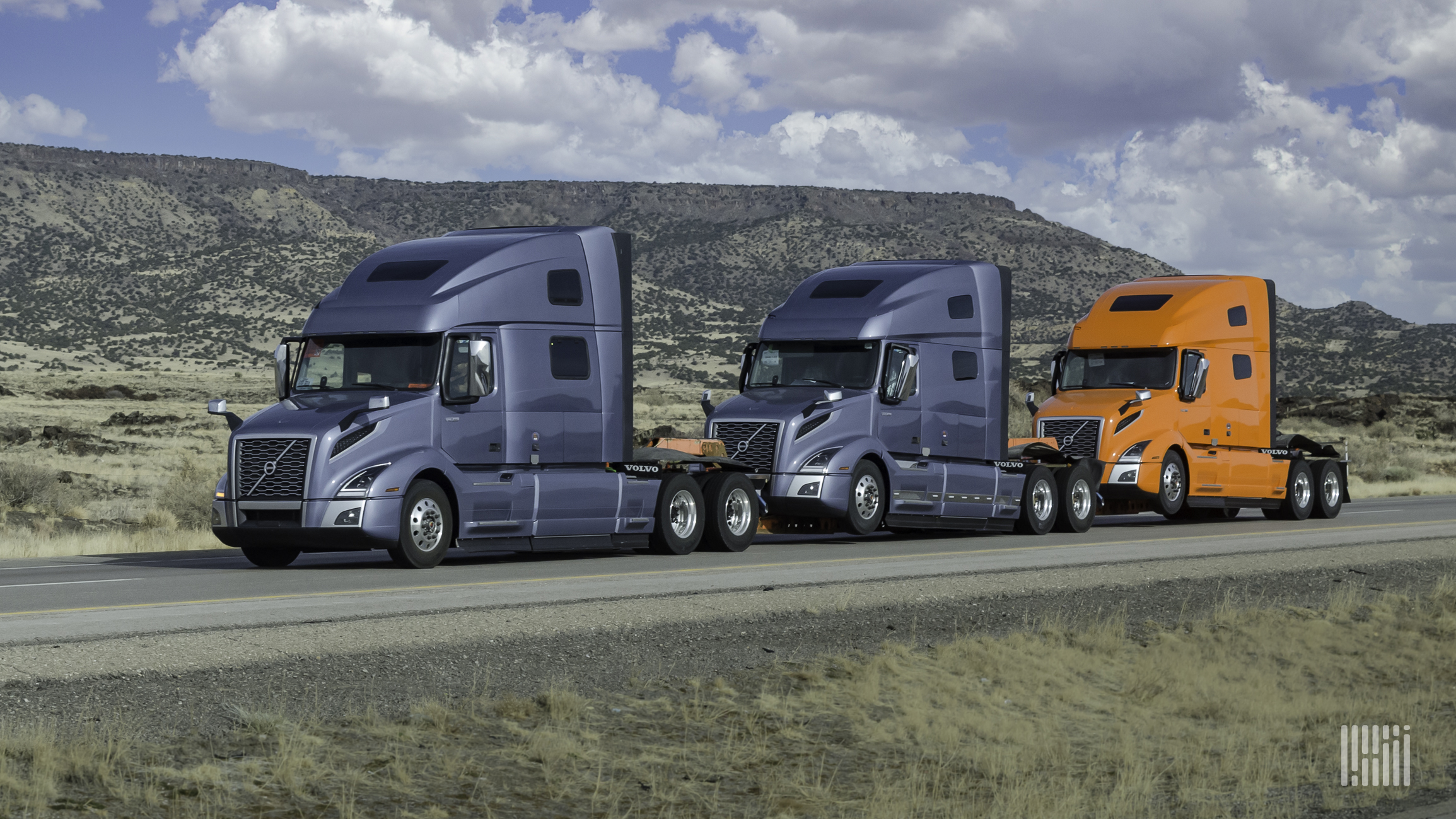Two blue and one orange Volvo sleeper cabs on a highway