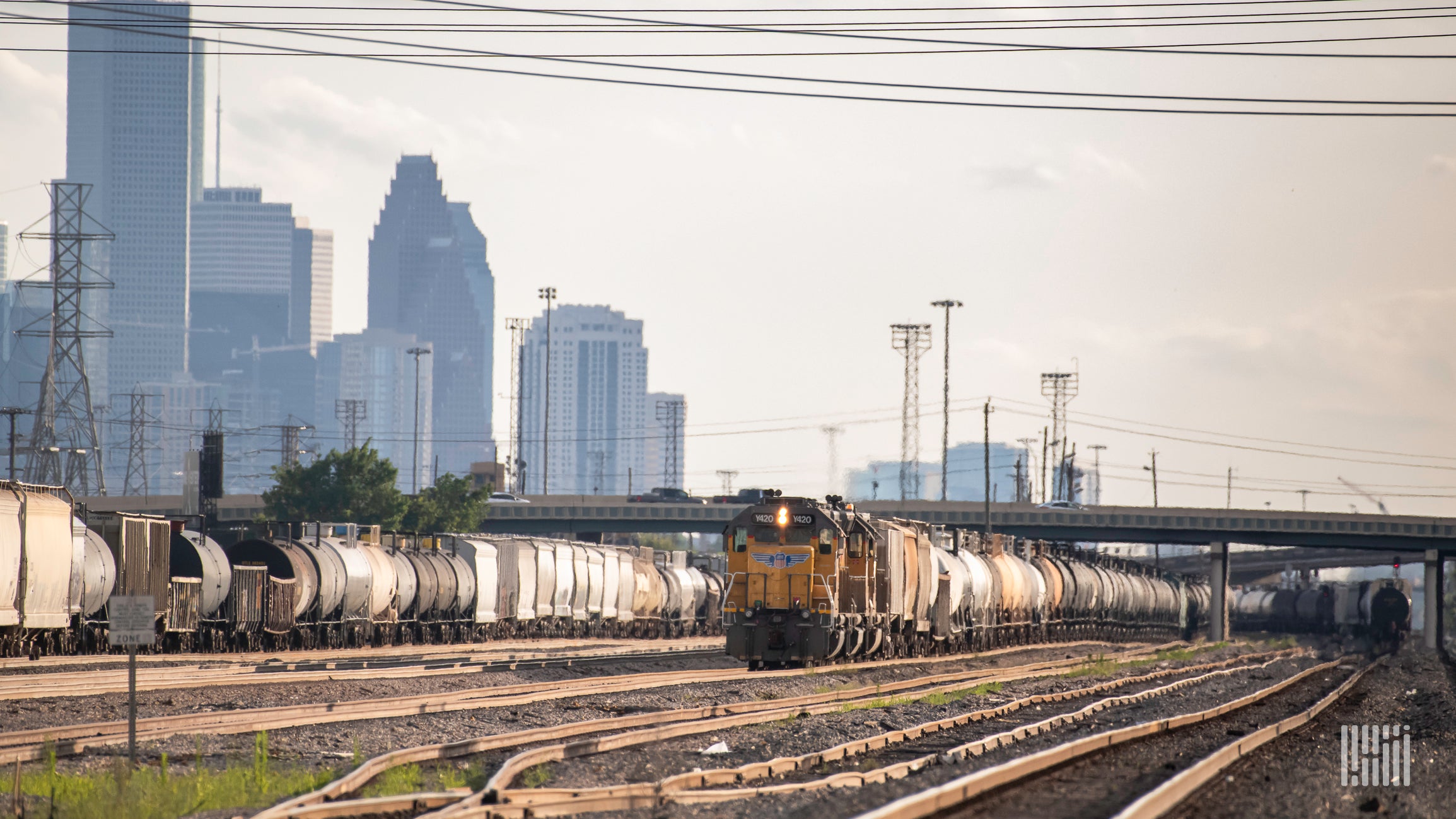 Two freight trains sit on train tracks in a rail yard. City skyscrapers are in the background.