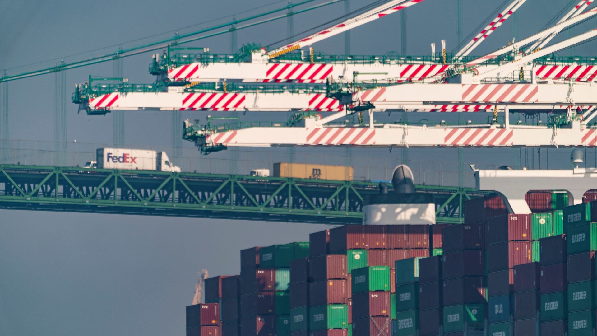 Giant ship cranes extend over containers stacked on a ship with a FedEx truck on a bridge in the background.
