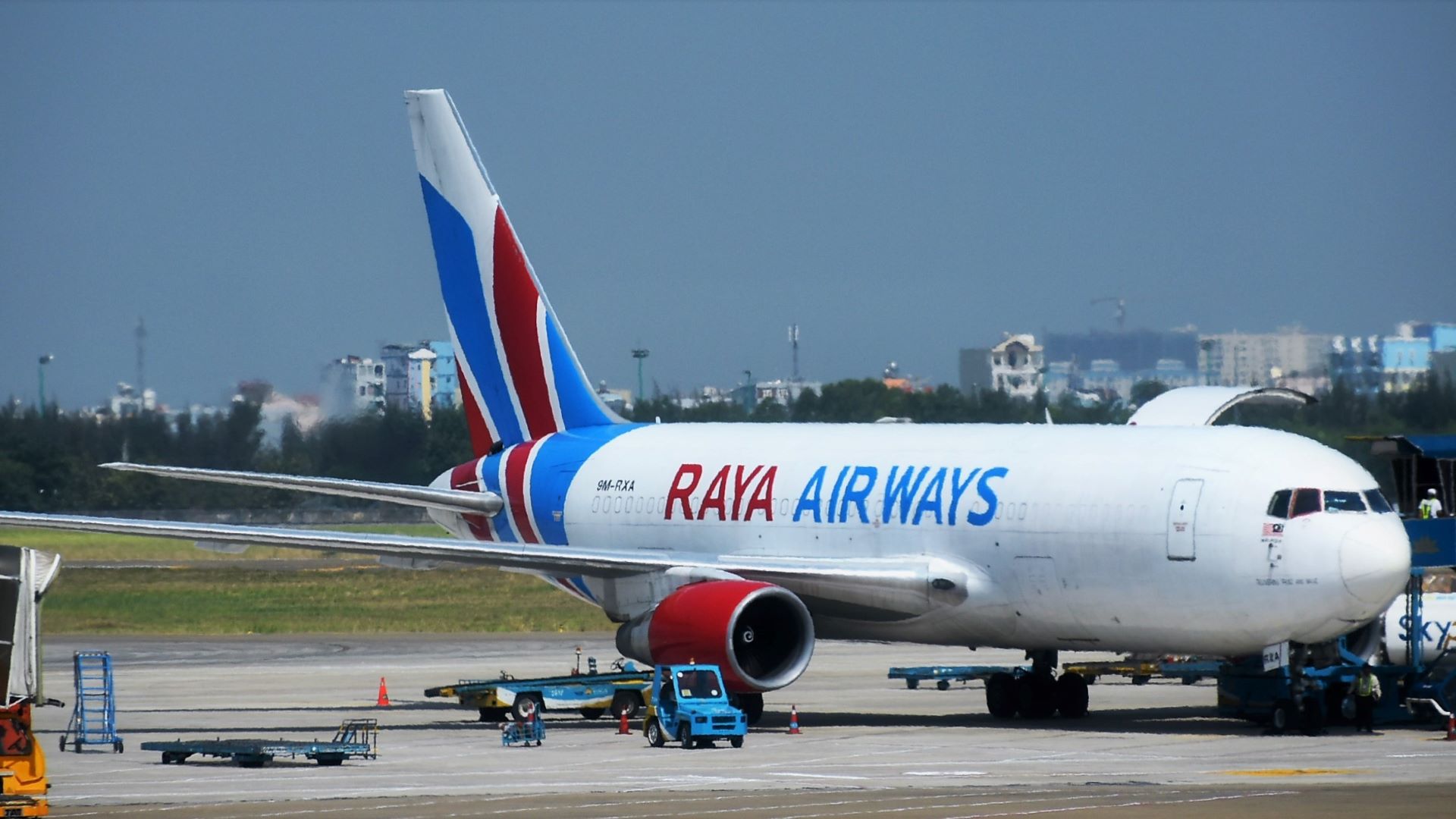 A white jetliner with blue/red tail and logo Raya Airways sits on the tarmac on a sunny day.