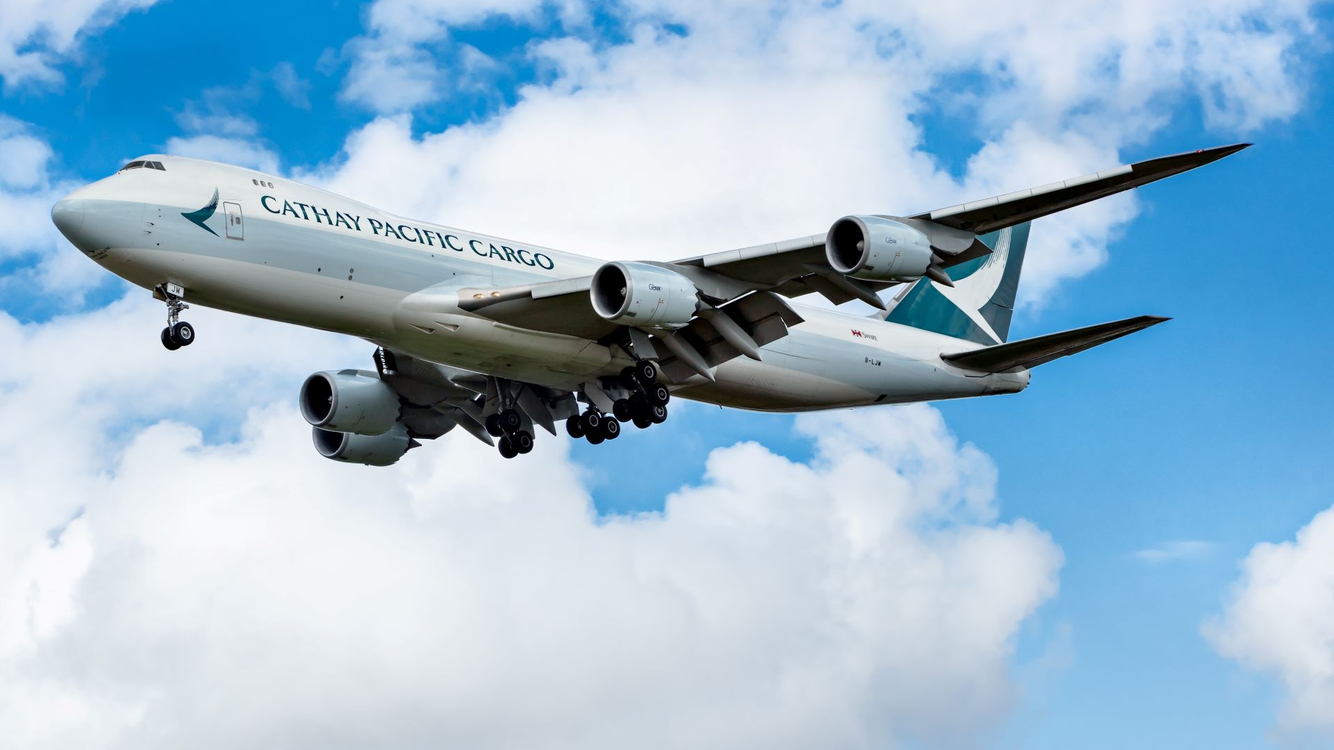 A white Cathay Pacific Cargo jumbo jet with teal tail viewed from below against blue sky as it comes in for a landing.