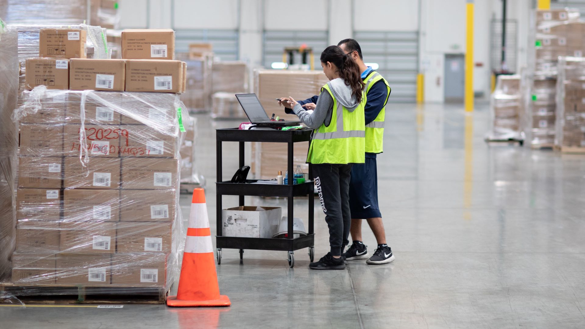Workers with yellow vests checking inventory information in a warehouse with boxes stacked up on foor.