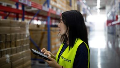 A woman with dark hair enters info into a digital pad while in warehouse. She is wearing a yellow safety vest.