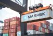 A crane lowers a large ocean container at a port with stacks of containers in the background.