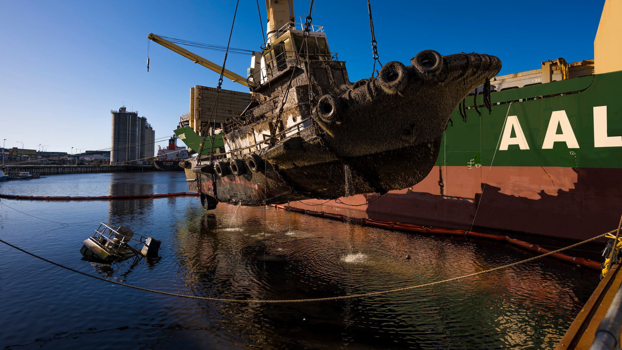 A mud-covered tugboat being lifted by cranes from a salvage vessel.