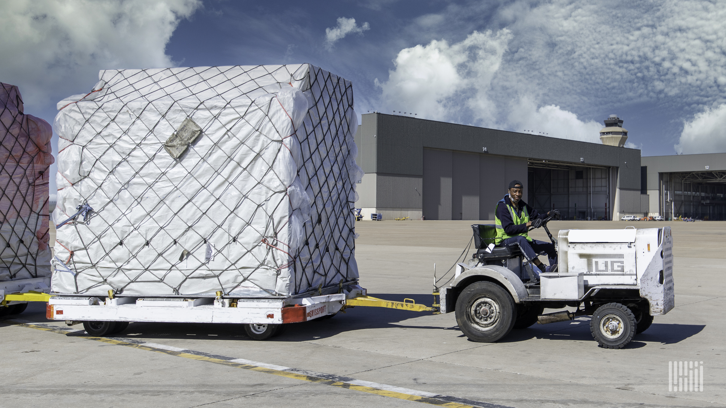 A large air freight container on a flat cart pulled by a tug.