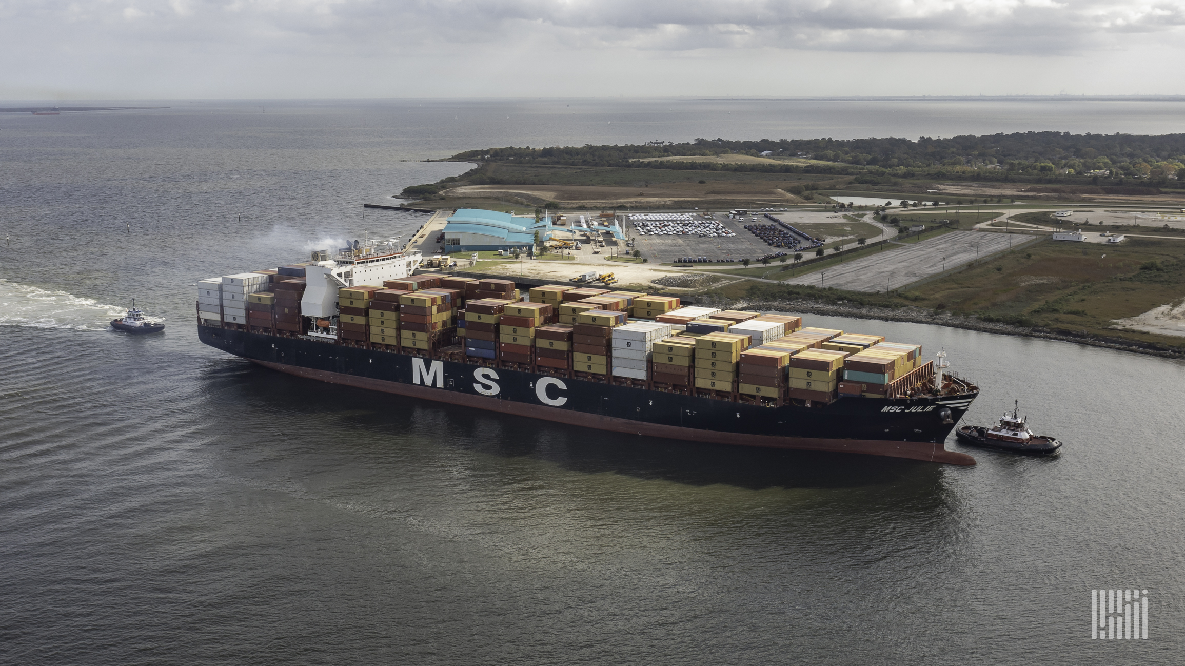 A massive container ship with MSC logo is pulled by tugs into the ship channel.