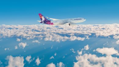 A colorful Hawaiian Airlines jetliner flying above white clouds with a blue sky in the background.