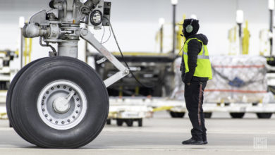 An airport ramp worker evaluates the wheel of a large cargo jet with a pallet of freight in the background.