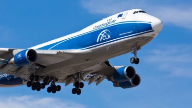 A blue-and-white jumbo jet for cargo comes in for landing against blue sky, looking up at plane.