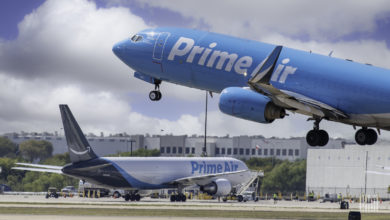 A baby-blue Amazon jet takes off with a white Amazon jet with dark blue tail sits in background.