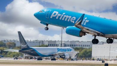 A baby-blue Amazon jet takes off with a white Amazon jet with dark blue tail sits in background.