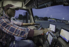 Man sits behind the steering wheel in the cab of a semi truck, touching the screen of a tablet on the dashboard