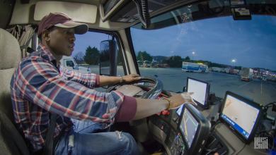 Man sits behind the steering wheel in the cab of a semi truck, touching the screen of a tablet on the dashboard