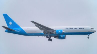 A white cargo jet, Maersk Air Cargo, with light blue tail flying with wheels down against gray sky.