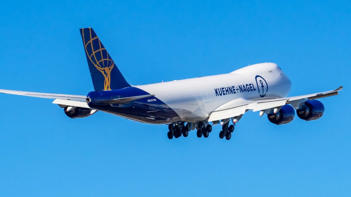 A white jumbo jet with a blue tail and lettering for Kuehne+Nagel takes off into the blue sky, viewed from the rear.