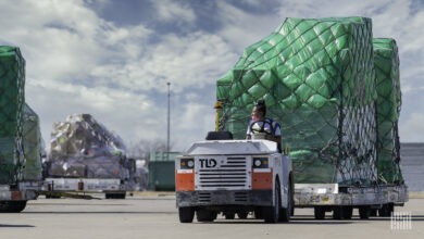 An airport cargo tug with a woman driver with black hair pulls a pallet with green shrink wrap on a bright day.