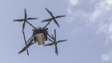A quad-rotor drone holding a small package in the air on a sunny day.